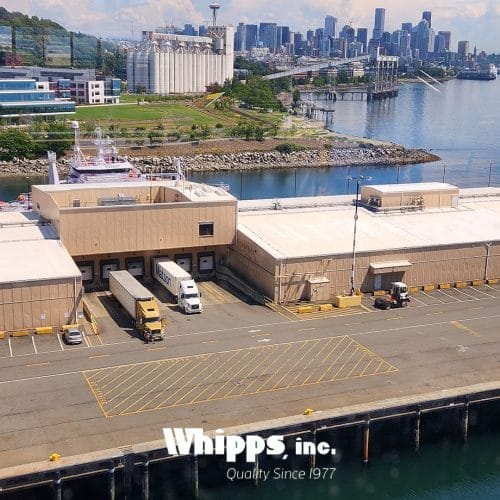 A dock with trucks and shipping facilities alongside a canal, with a city skyline and green hills in the background.