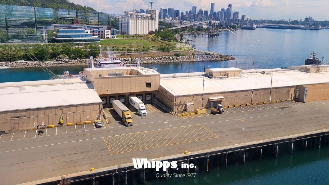 A dock with trucks and shipping facilities alongside a canal, with a city skyline and green hills in the background.