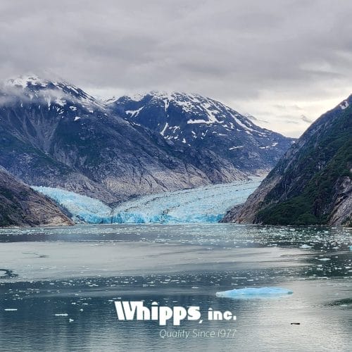 A glacier flowing into a calm body of water surrounded by rugged mountains, with floating icebergs scattered across the surface.