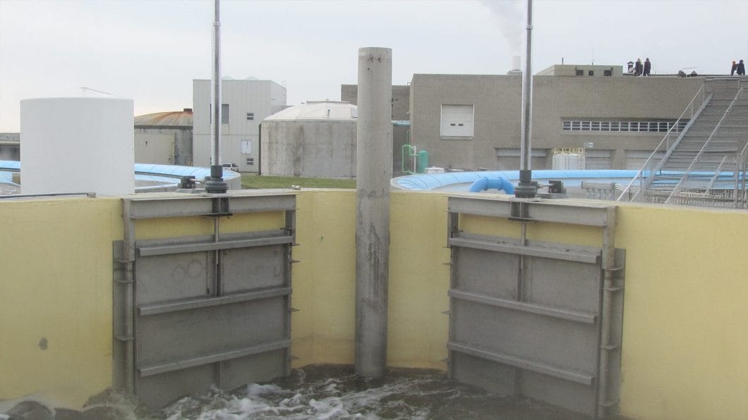 A pair of stainless steel slide gates mounted on a yellow concrete wall, controlling water flow at an industrial water management facility.