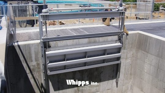 A stainless steel weir slide gate manufactured by Whipps, Inc., installed in a concrete water control structure. The gate is mounted on a vertical track and is designed to open downward for efficient water regulation in limited overhead space. The Whipps, Inc. logo is visible at the bottom of the image.
